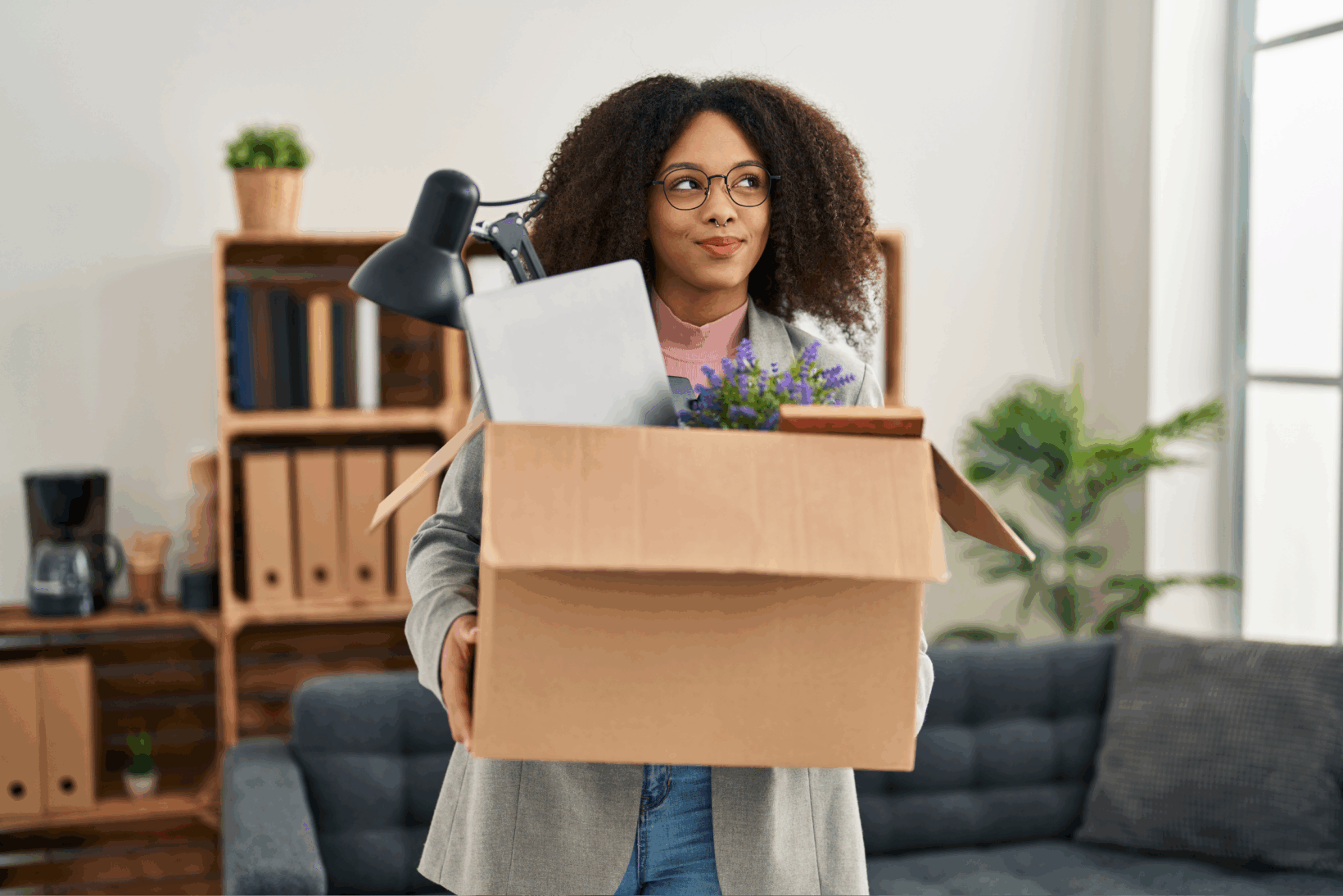 A young woman holding a box with a lamp, a folder, and a plant in an office